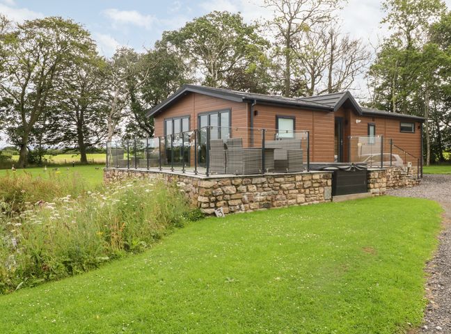 A single-story cabin with a glass railing patio and outdoor furniture surrounded by grass and trees at Henry Haven Nateby near Garstang