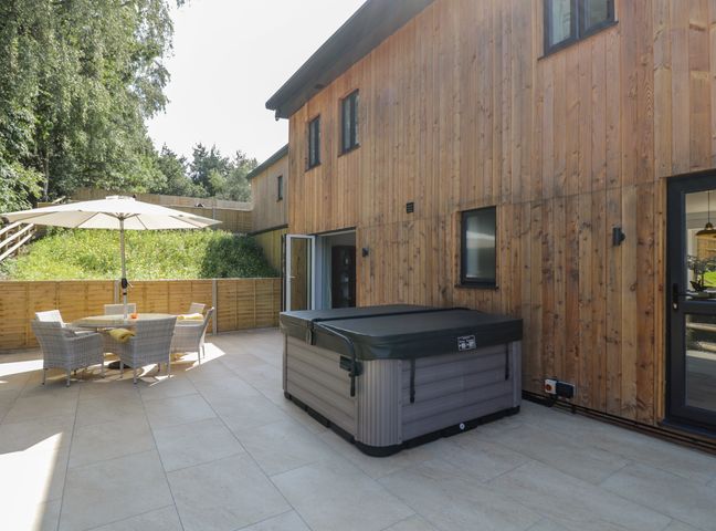 An outdoor patio with a hot tub wood siding on the house a table with chairs and an umbrella at Stag Cottage in Lytchett Matravers