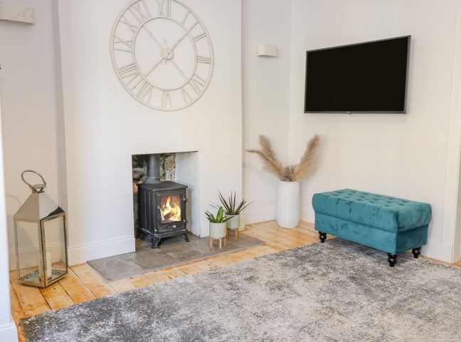 A room with a small fireplace with fire a large wall clock a teal bench a wall-mounted television and plants at The Lazy Seal in Spittal