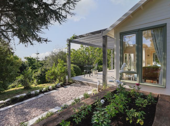A garden with flowers next to a gravel path and a wooden porch with table and chairs at Meadow Lodge @ Seawardstone in Blackawton