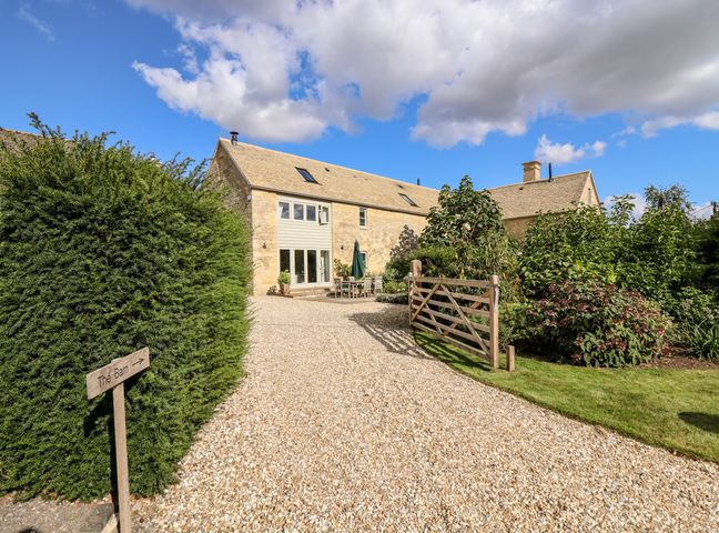 A stone barn with a gravel driveway a wooden fence and garden plants at Stow Cottage Barn in Churchill Oxfordshire