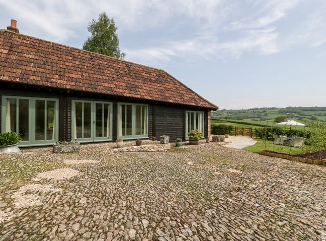 An exterior view of a rustic building with tiled roof and multiple glass doors opening onto a cobblestone courtyard with potted plants and a garden area at The Shippon in Honiton