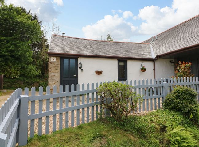 Exterior of a small white house with grey door and window behind a grey picket fence with greenery in front at Ash Cottage in Combe Martin