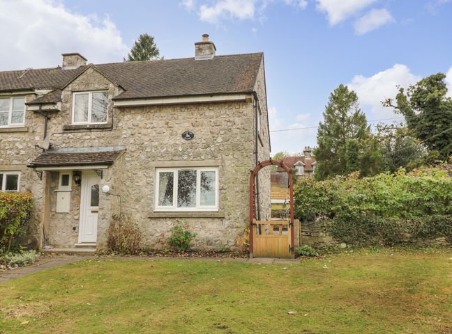 A stone cottage with a white door and window and a wooden gate in front of grass at Cloud View Cottage in Thorpe near Ashbourne