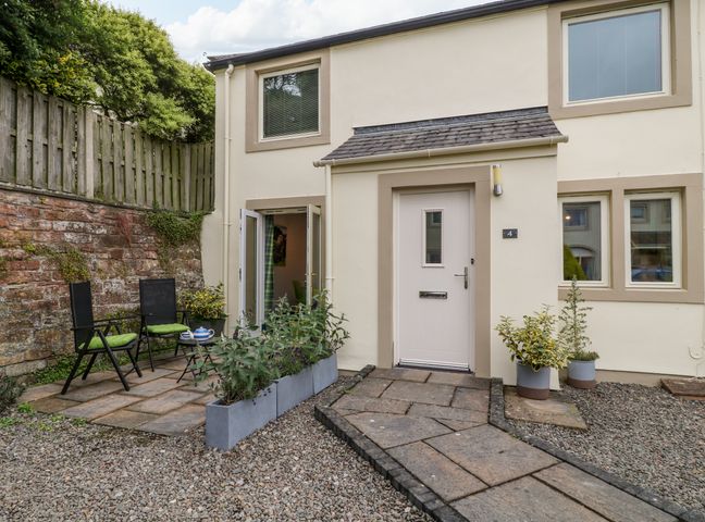 A paved patio with two black chairs and a small table with a teapot outside a cream building with windows and a door at Herdwick Cottage in Brampton Cumbria