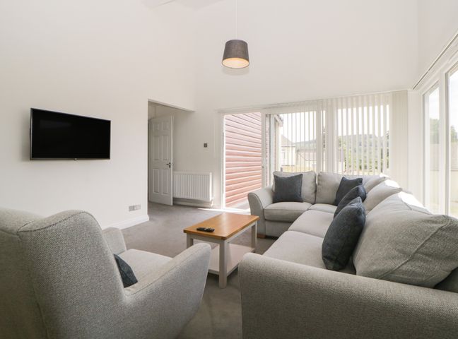 A living room with a wall-mounted television gray sofas a wooden coffee table and large windows with vertical blinds at Harcombe House Bungalow 5 in Chudleigh