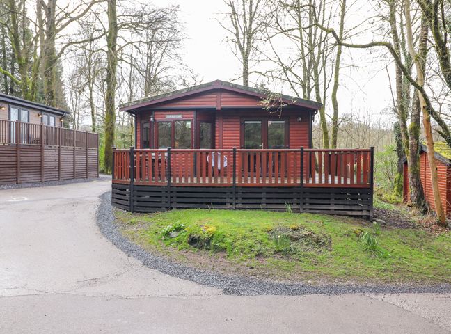 A wooden cabin with a fenced porch surrounded by trees at Alfresco Lodge in Skiptory Howe 37B White Cross Bay near Windermere