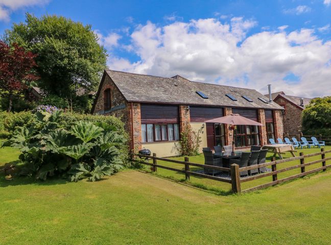 A stone barn with outdoor dining table umbrella and lawn chairs on a grass lawn at Apple Barn in South Molton