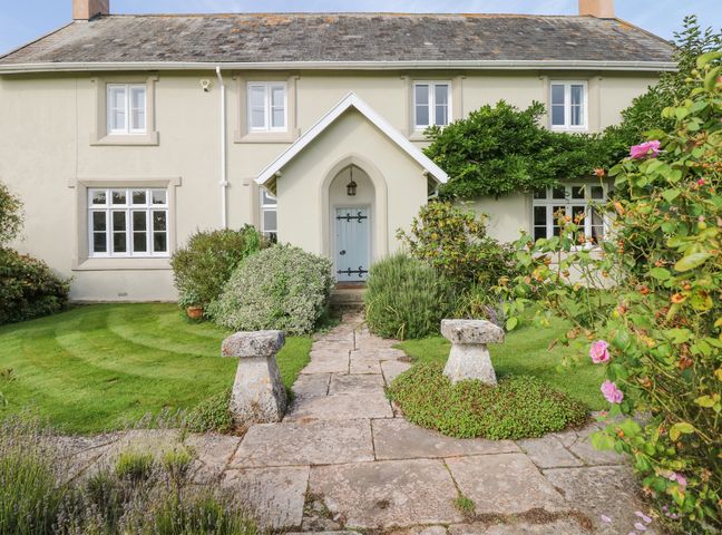The front exterior of a house with a stone pathway leading to a blue front door surrounded by bushes and flowers at Crablake Farmhouse in Exminster