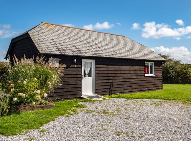 A single story black wooden building with a white door and window surrounded by grass and gravel at Puffin Lodge in Port Isaac