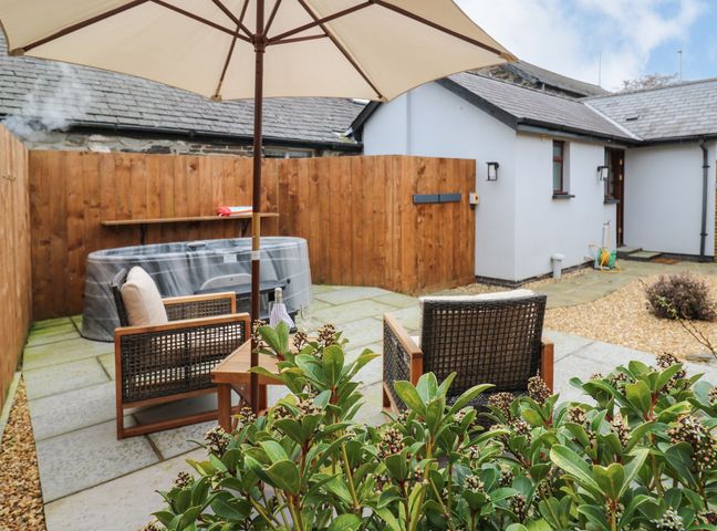An outdoor patio area with a hot tub wooden chairs a table with an umbrella and a wooden fence at Llygad Y Dydd in Bow Street Wales