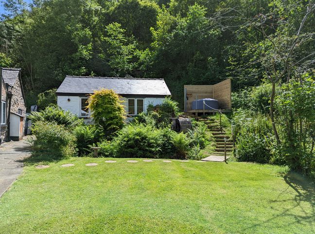 A small white cottage with a garden and wooden steps leading to a deck with a hot tub surrounded by trees and bushes at Little Pandy Cottage in Afonwen near Caerwys