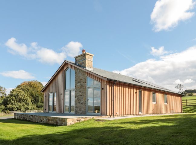 A wooden house with large windows and a stone chimney on a grassy lawn at Clover Cottage in Corbridge