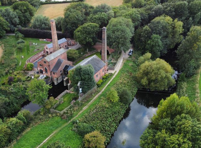 A rural scene with brick buildings and tall chimneys near a river surrounded by trees and greenery at The Pump House Forge in Misterton