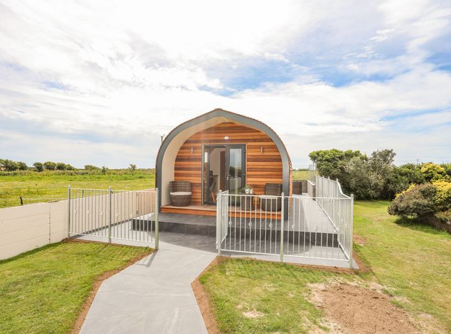 An arched wooden cabin with two chairs on the porch and a fenced patio in a grassy area at Caban Cariad in Bryngwran near Bodedern