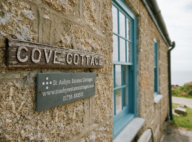 A stone wall of a cottage with a wooden sign reading Cove Cottage and a grey plaque with contact information at Cove Cottage in Porthgwarra