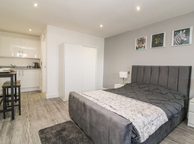 A bedroom with a gray upholstered bed a white wardrobe a small kitchen area with cabinets a wooden table and stools and framed animal prints on the wall at Chalet 3 in Sherburn