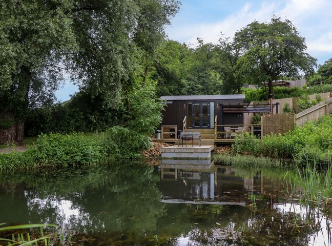 A small cabin with outdoor seating overlooking a pond surrounded by trees and greenery at The Shepherds Hut at Bridge Lake Farm & Fishery in Chacombe