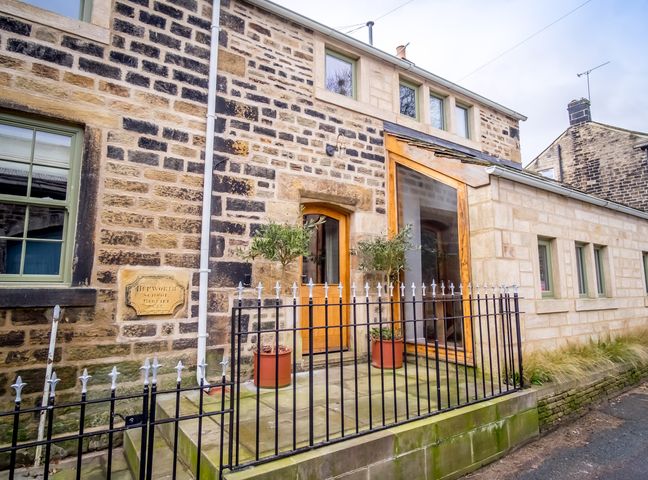 Exterior view of a stone building with windows wooden door stone steps and potted plants behind a black metal fence at School Cottage in Hepworth near Holmfirth