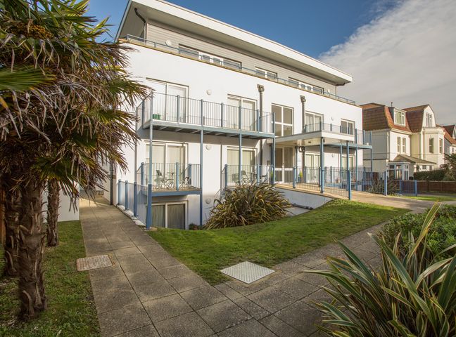 The exterior of a modern white apartment building with balconies and a pathway surrounded by grass and plants at 1 Blue Waters in Southbourne