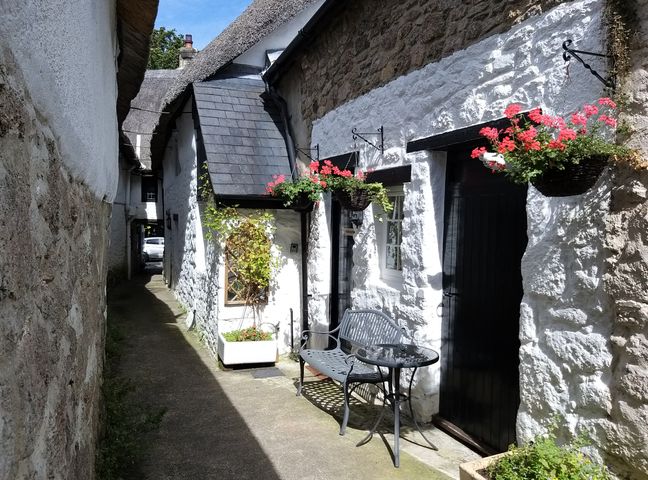 A narrow outdoor passage with white stone walls a bench and table and hanging flower baskets at Fox Hat Cottage in Chagford