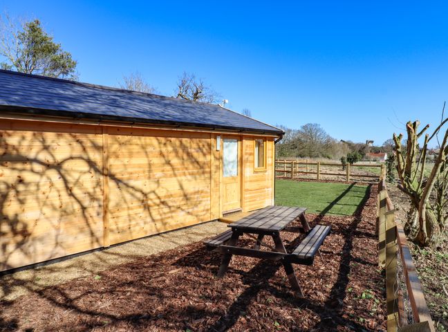 An outdoor area with a wooden picnic table beside a wooden building and a fenced grassy area at Buttercup Lodge in Theberton near Leiston
