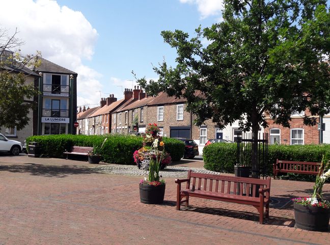 A small urban park with benches flower pots a tree and residential buildings in the background at No.37 in Beverley