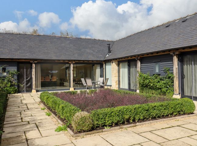 An outdoor courtyard with a square garden bed surrounded by hedges and patio furniture at Hawthorn Barn in Northleach