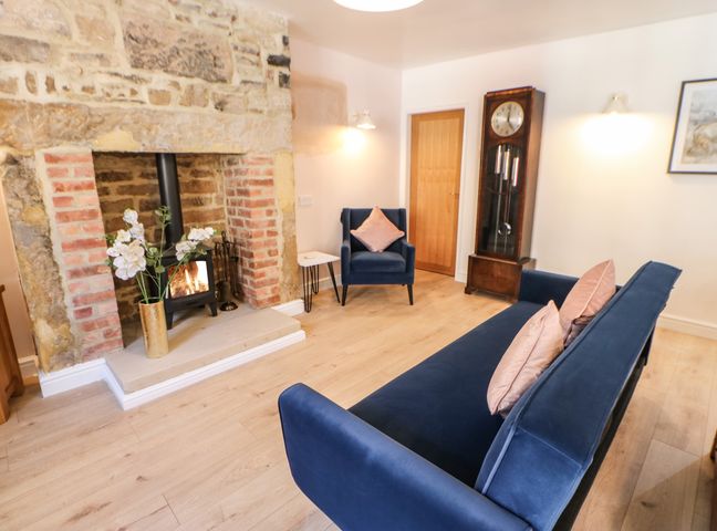 A living room with a stone fireplace and wood stove a blue armchair and sofa with pink cushions and a grandfather clock at 3 Mill Wynd in Staindrop