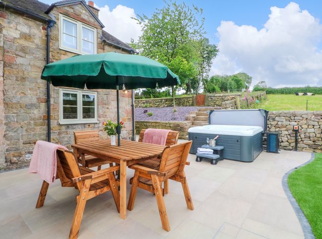 An outdoor patio with a wooden table and four chairs under a green umbrella next to a hot tub at Small Lane Cottage in Blackwood Hill near Endon