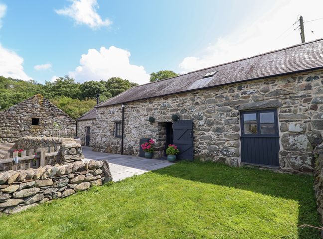 A stone cottage with slate roof and black doors and windows with potted plants on a paved area and a stone wall in the garden at Beudy Bach in Llanfair near Harlech