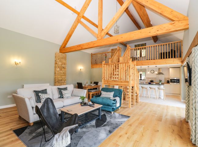 An open living space with wooden beams a staircase leading to a loft kitchen with bar stools and a seating area with sofas and a lounge chair at Petals Patch in Lower Slaughter