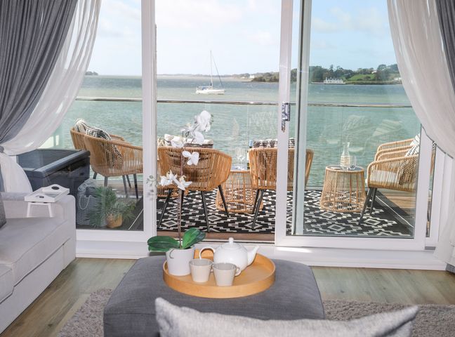 A living room with a teapot and cups on a tray on an ottoman facing a balcony with wicker chairs and a view of water at Seaclusion in Y Felinheli