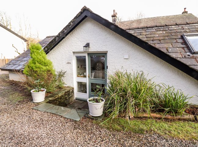 An exterior entrance to a white building with a sloped roof surrounded by plants and gravel at Peony Cottage Lakeside near Newby Bridge