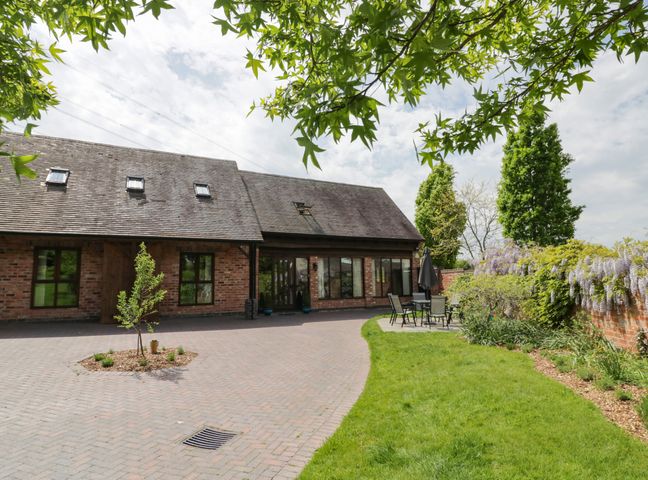 A brick building with multiple windows and skylights with a paved driveway and a small garden area with trees and patio furniture at The Stables in Belbroughton