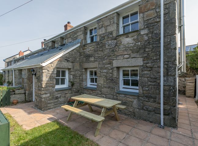 An outdoor patio with a wooden picnic table next to a stone cottage at 15 Boscaswell Downs in Pendeen