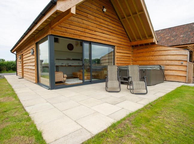 An outdoor patio with two reclining chairs and a hot tub next to a wooden house with glass sliding doors at Starling Lodge in Wedmore