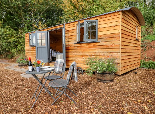 A small wooden cabin with open doors and windows a table with two chairs and a bottle of wine outside at Hideaway Cabin in Cadbury near Thorverton
