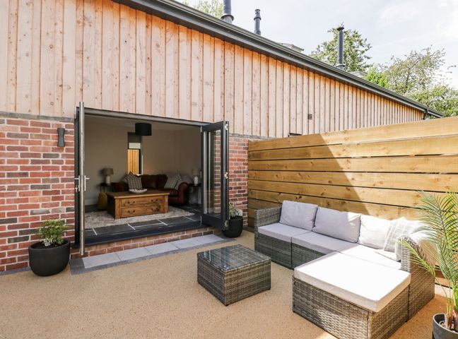 An outdoor patio area with gray cushioned wicker seating and a glass table next to a building with wooden and brick walls at Little Lugg in Cross Keys near Hereford