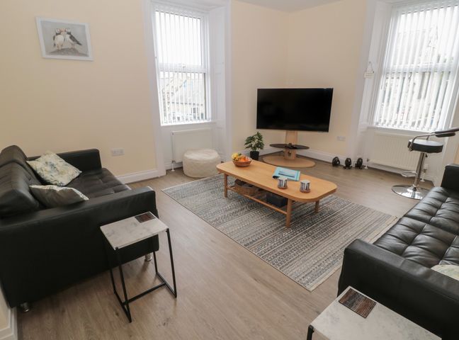 A living room with two black sofas a wooden coffee table with mugs and a fruit bowl a flat screen tv on a stand a chair by the window and a small plant at Amble Watch in Amble