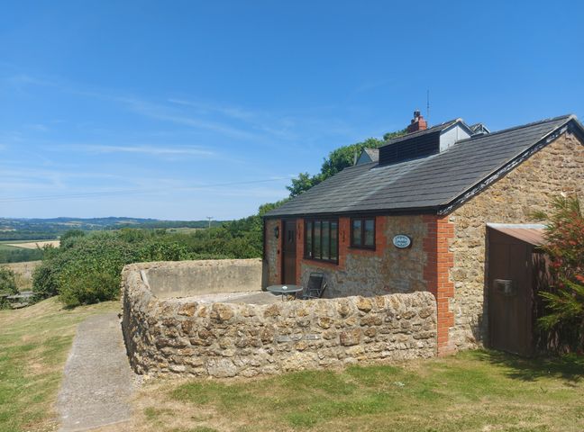 A small stone building with a fenced patio and a table with chairs in a rural setting at The Sty in Bridport