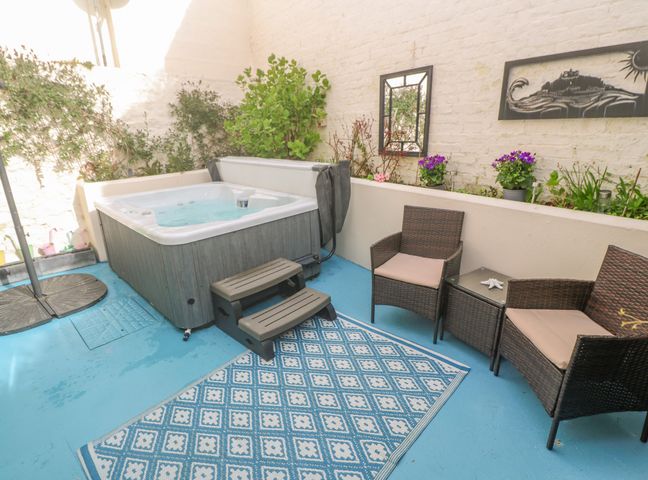 An outdoor patio area with a hot tub two wicker chairs a side table a patterned rug and potted plants on a planter at Driftwood in Marazion