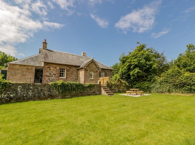 A stone cottage with chimney and steps leading to a wooden gate in a grassy yard with table and benches surrounded by trees and bushes at North Firbank - Culzean Castle in Maybole