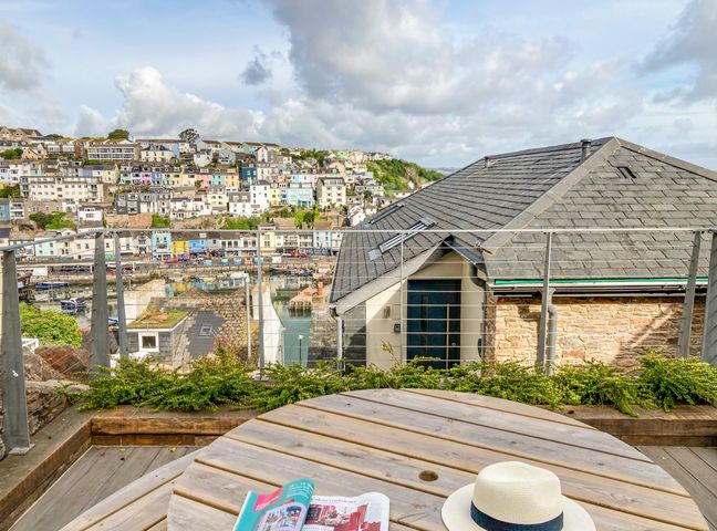A wooden outdoor table with a magazine and a hat on a balcony overlooking a coastal town with multi-colored houses at Bay View in Brixham