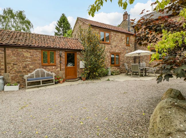 A stone house with a gravel courtyard a wooden bench flower pots and an outdoor dining table with an umbrella at Sutton Barn in Hope Mansell