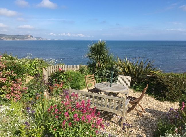 A garden with a wooden table and chairs overlooking the sea with flowers and greenery at 4 East Cliff in Lyme Regis