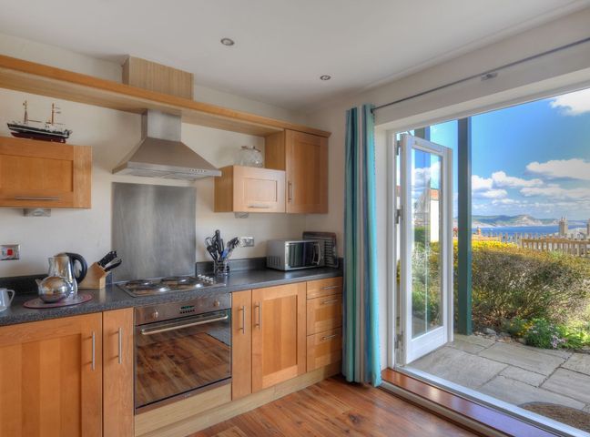 A kitchen with wooden cabinets, a stove, microwave, kettle, knife block, and an open door leading to a garden with a sea view at 8 Buckfields Lyme Regis