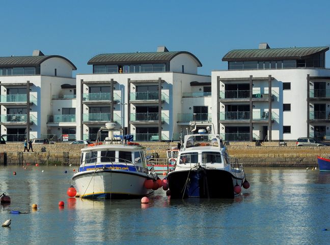Two boats moored in a harbor with modern apartment buildings and parked cars in the background at Harbour View in West Bay