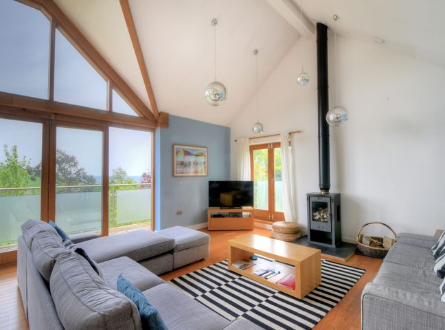 A living room with grey sofas a coffee table a wood stove and large windows at Pippin in Charmouth