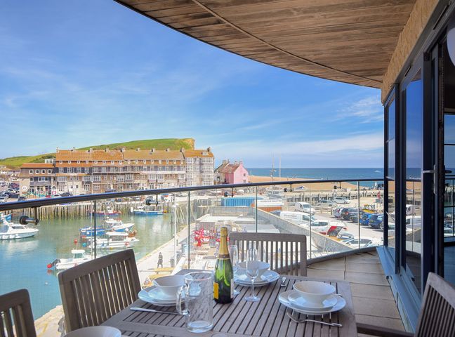 A balcony with a wooden dining table and chairs overlooking a harbor with boats and buildings at Venezuela West Bay